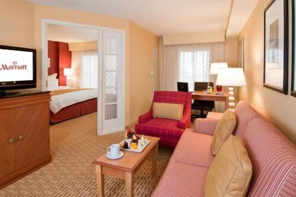 A hotel room with a bed, a red armchair, a sofa, a desk with a lamp, and a television.