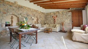 A rustic living room with stone walls, wooden beams, a stone fireplace, a sofa, a dining table with chairs, and a wooden door. - Son Palanca, Finca 5starshome Mallorca (Sineu)