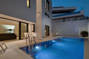 Un área de piscina exterior moderna con piscina cubierta por vidrio, sillas de descanso y una mesa de comedor.