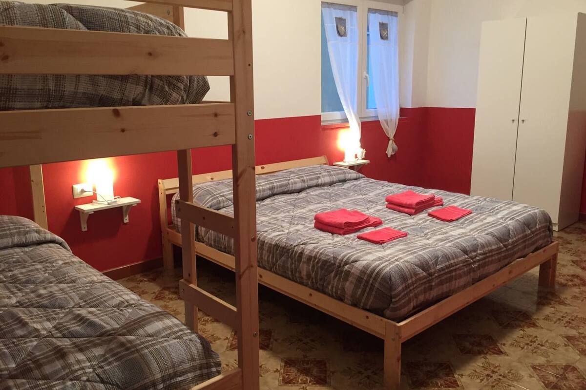 Habitación tipo dormitorio con dos literas, una cama individual, una mesita de noche con lámpara y una ventana con cortinas.