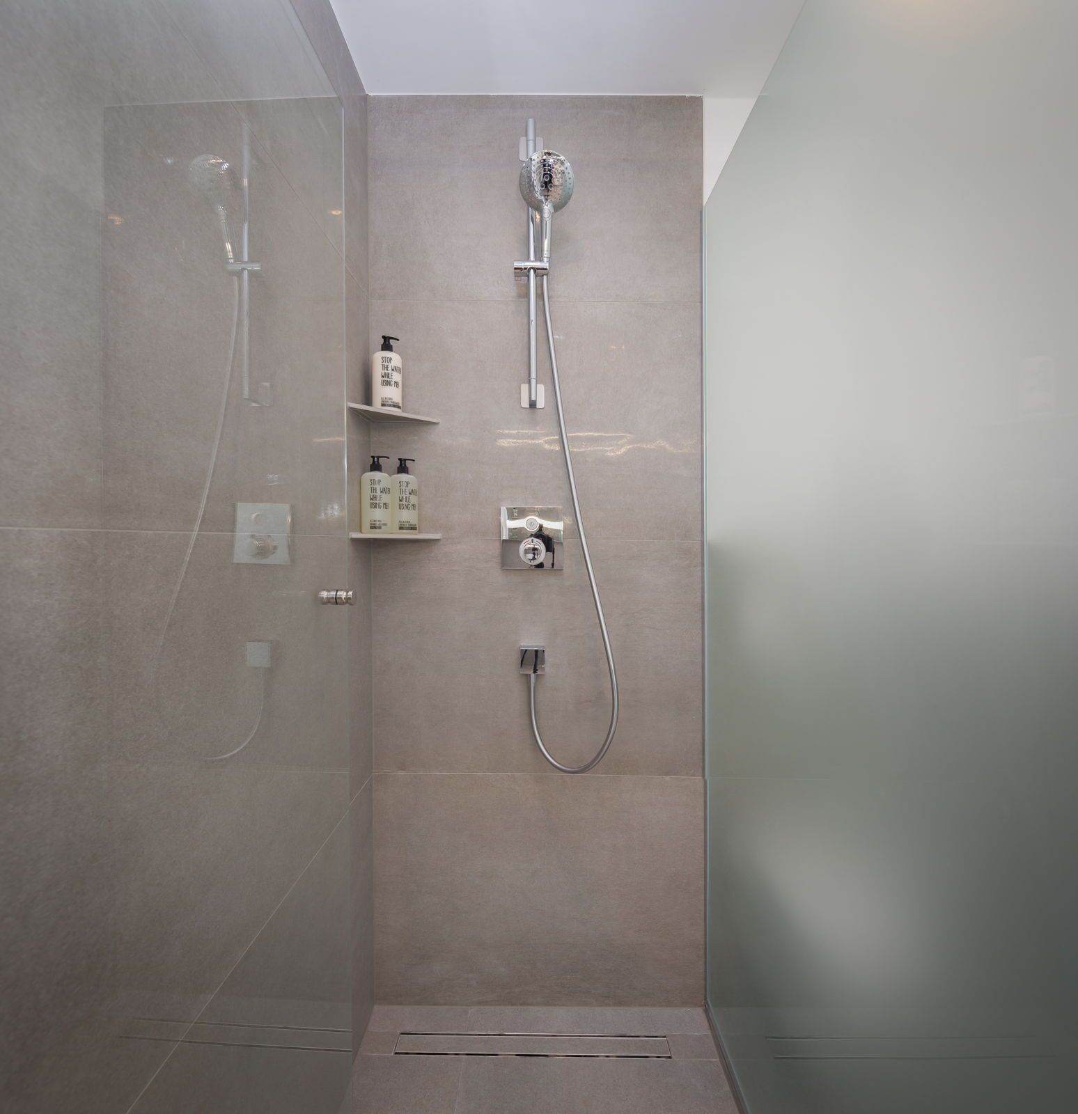 En moderne bruseniche med håndbruser og vandhane fastgjort på væggen. To små hylder med toiletartikler.