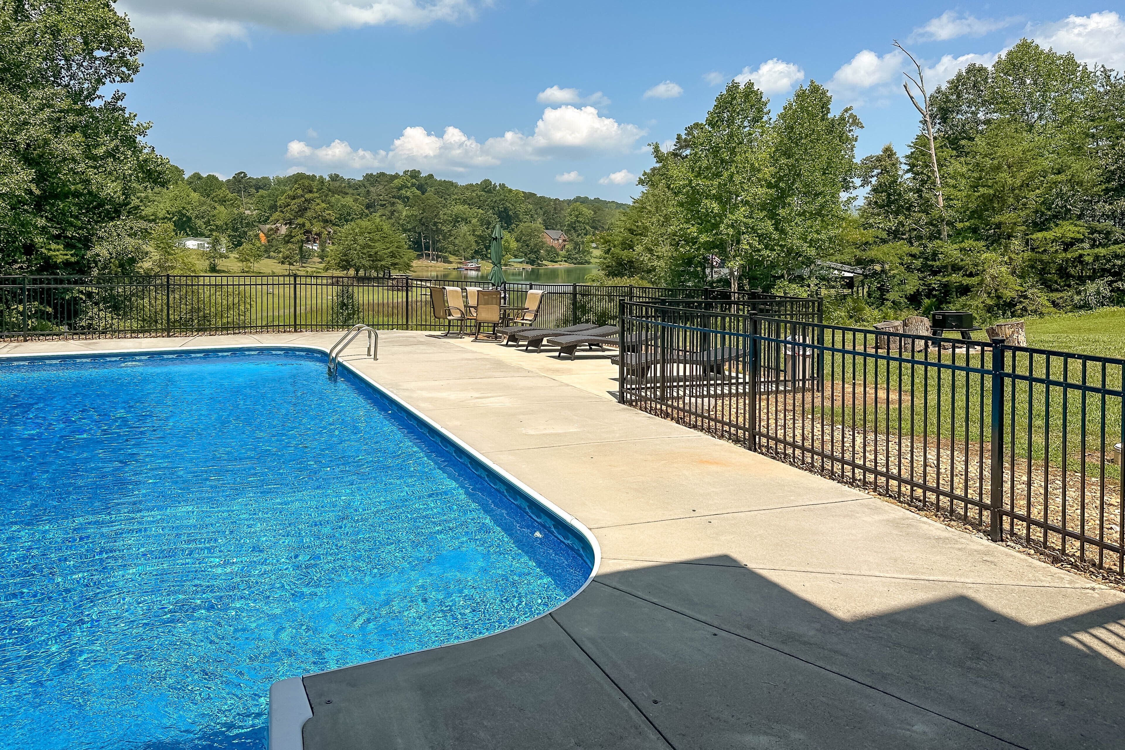 A swimming pool with a black metal fence, lounge chairs, and a view of a lake and forested hills.