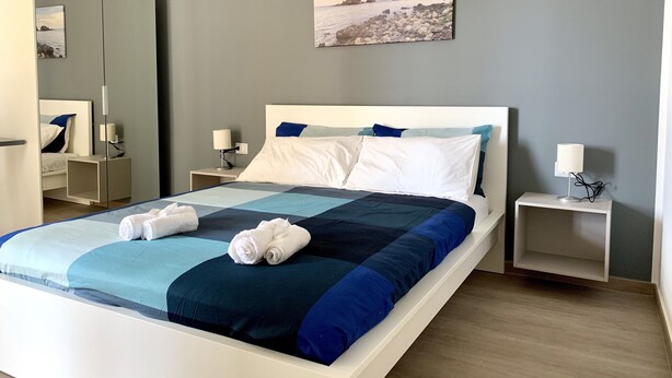 A neatly made bed with a blue and white quilt, two white pillows, and rolled towels. There are two bedside tables with lamps, a large mirror, and a framed picture on the wall. - Ambasciatori B&B (Catania)