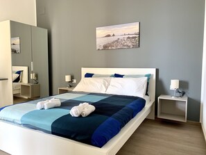 A neatly made bed with a blue and white quilt, two white pillows, and rolled towels. There are two bedside tables with lamps, a large mirror, and a framed picture on the wall. - Ambasciatori B&B (Catania)