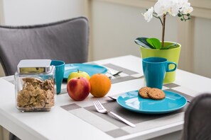 A breakfast table with a checkered tablecloth, a blue plate with cookies, a glass jar of cereal, a blue mug, a green plant in a yellow pot, and a fruit bowl with apples and an orange. - Ambasciatori B&B (Catania)