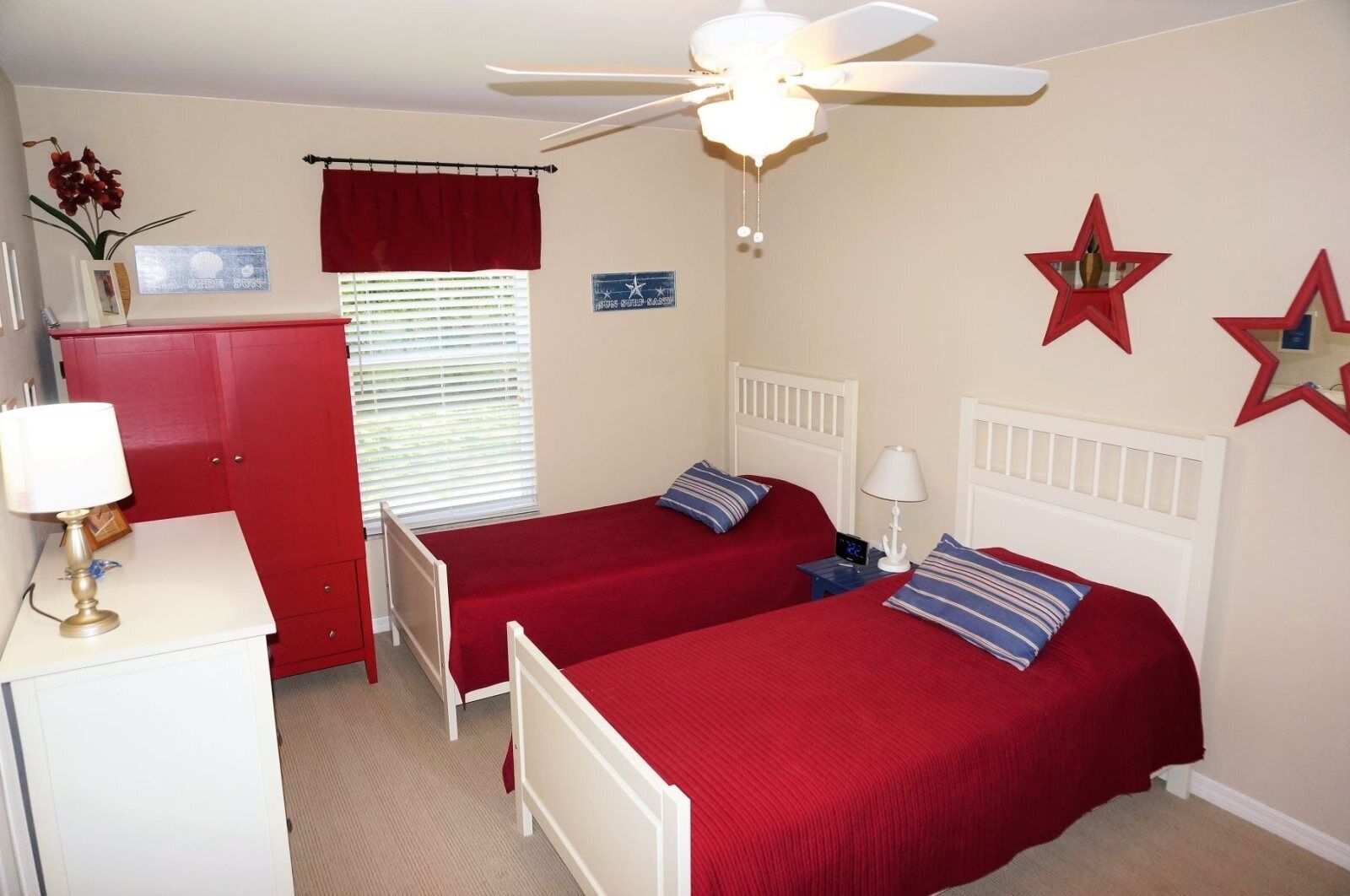 Une chambre avec deux lits simples, un ventilateur de plafond, une armoire rouge et une fenêtre avec des stores.