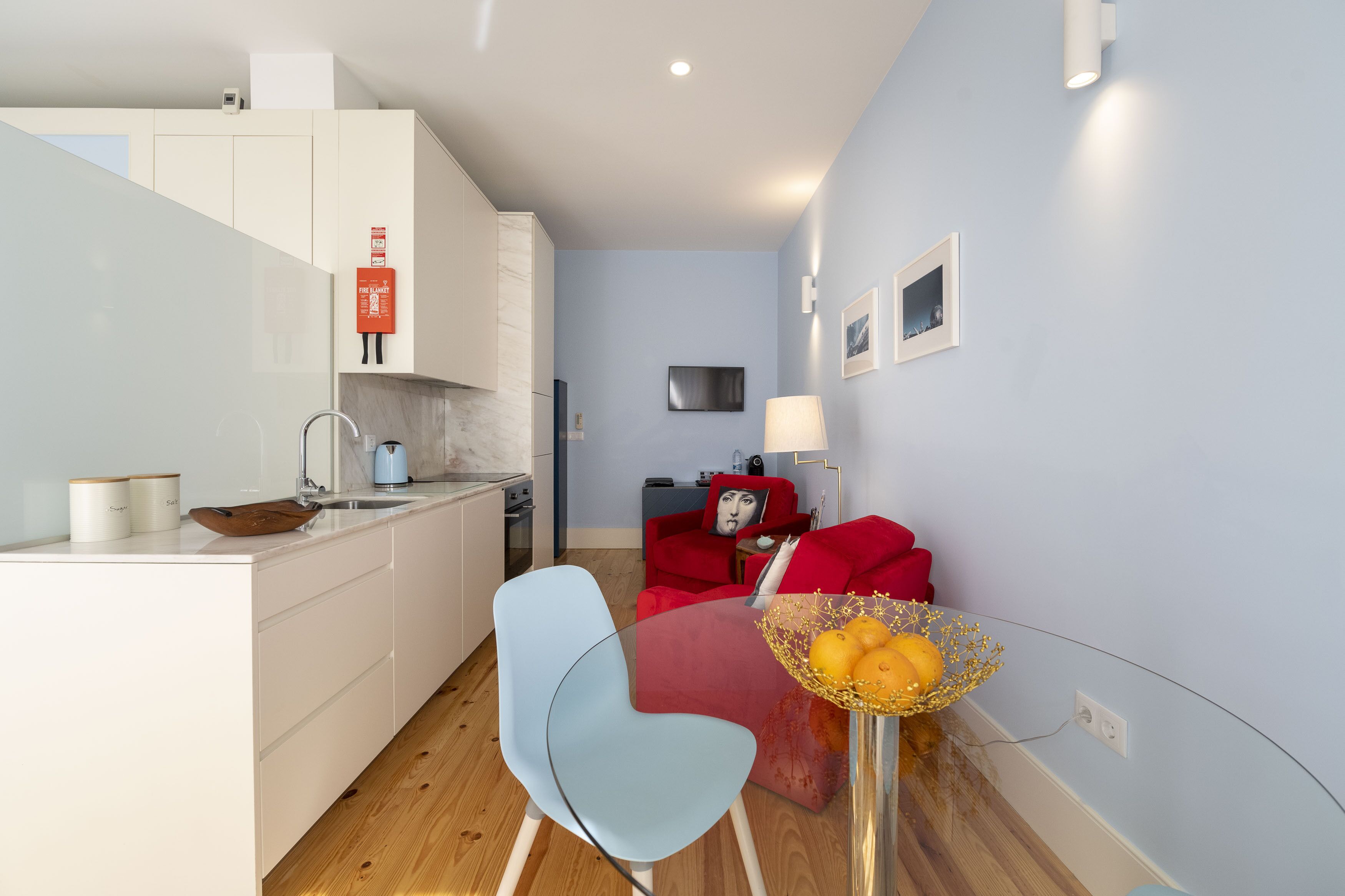 A modern kitchen with white cabinets, a red sofa, and a glass dining table with a fruit bowl.