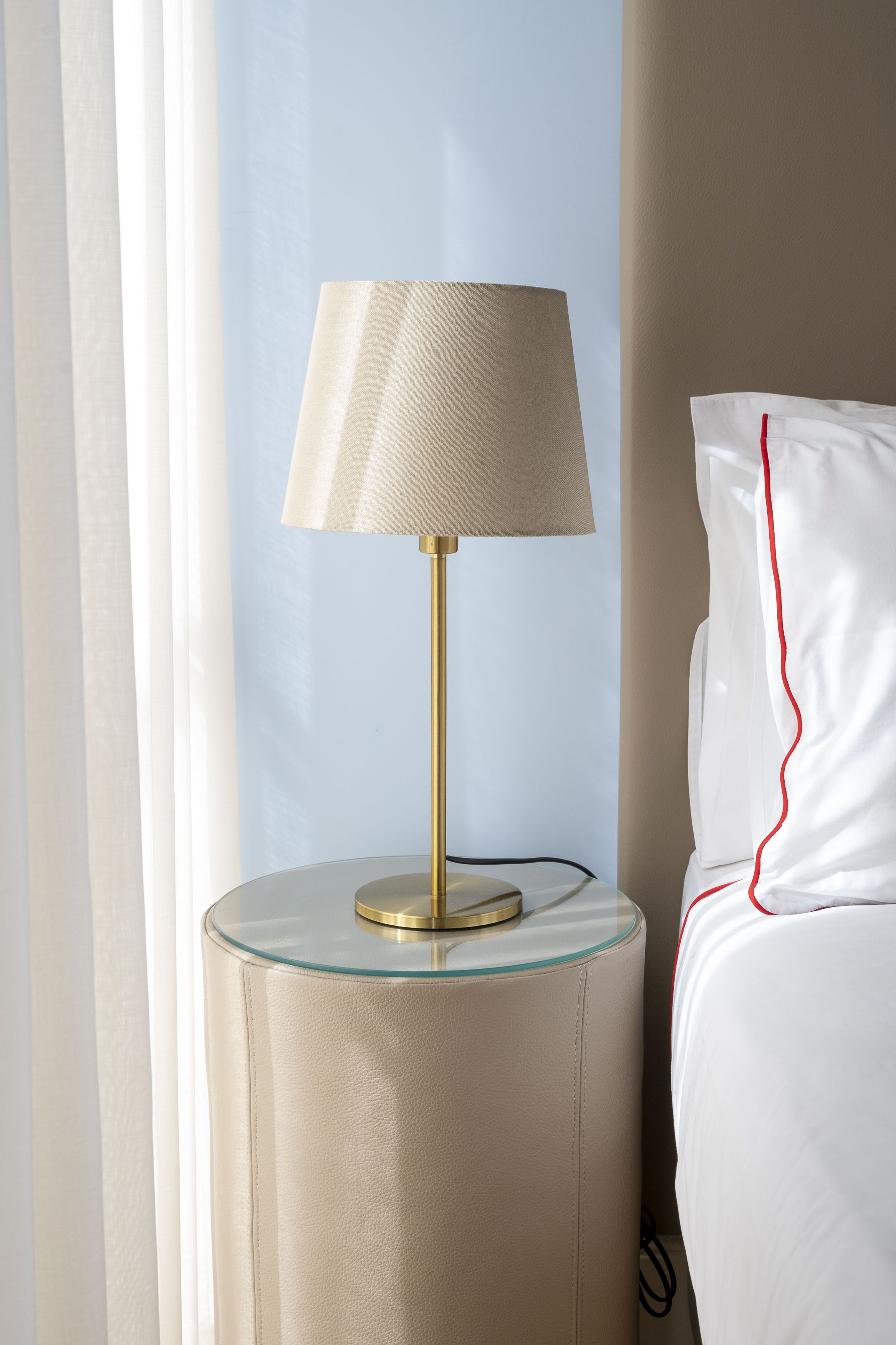 Un lit avec une literie blanche et une bande rouge décorative, une table ronde en verre avec un pied doré et une lampe de couleur beige.