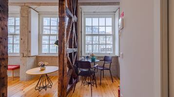 A room with wooden floors, a round table with chairs, a vase with flowers, and large windows with a view of buildings.
