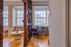 Een kamer met houten vloeren, een ronde tafel met stoelen, een vaas met bloemen en grote ramen met uitzicht op gebouwen.
