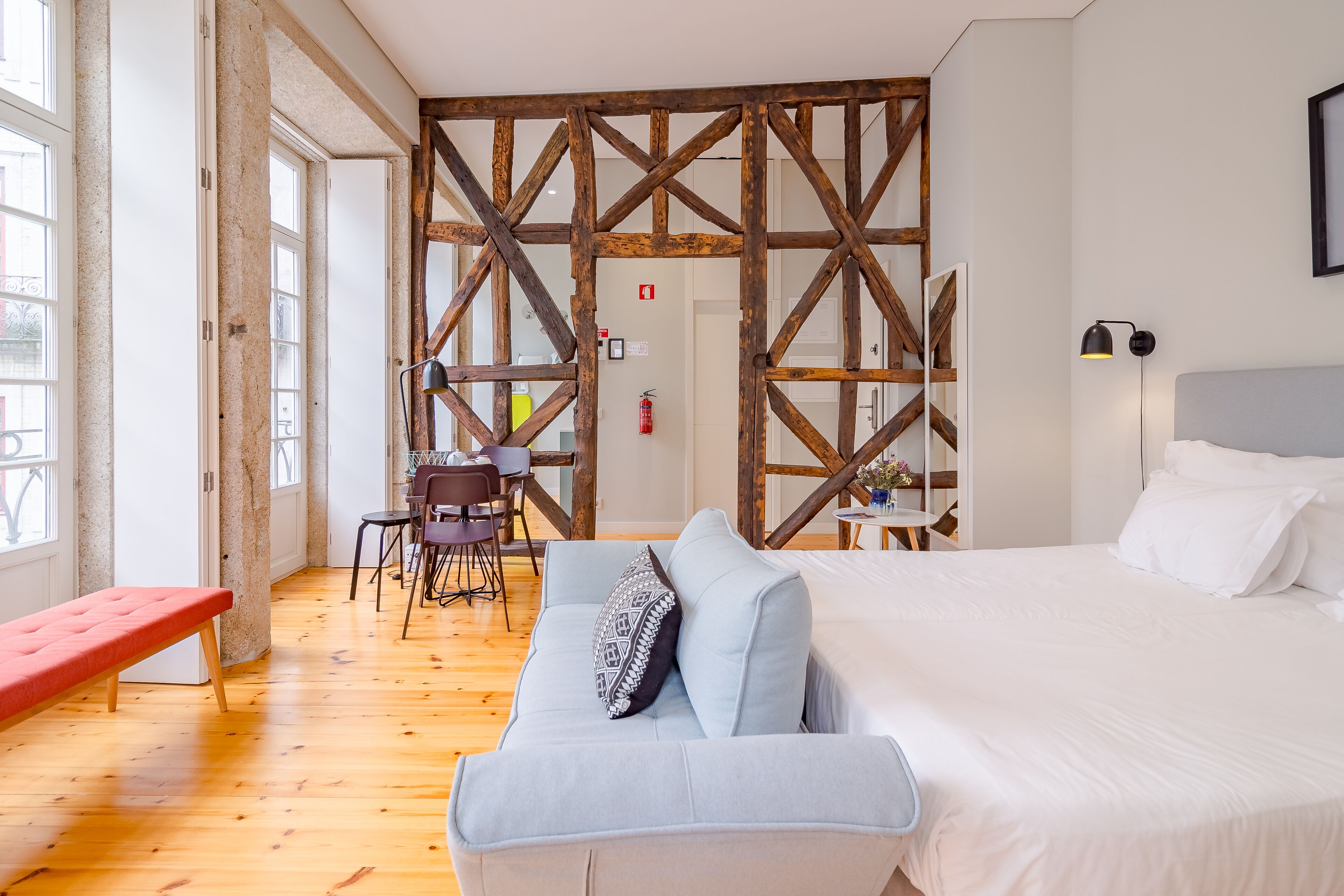 A hotel room with a wooden floor, a bed with white linens, a light blue sofa with a patterned pillow, a red bench, and a wooden partition wall.