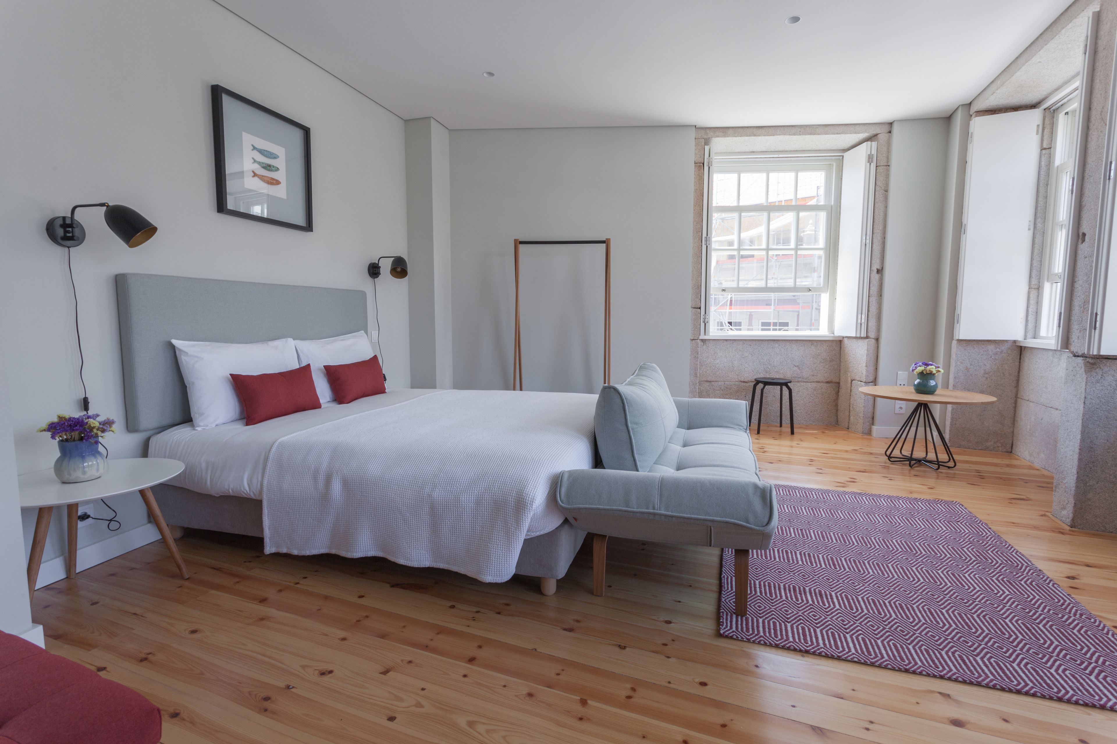 A modern bedroom with a large bed, a grey sofa, a small round table, and a window with a view of buildings.