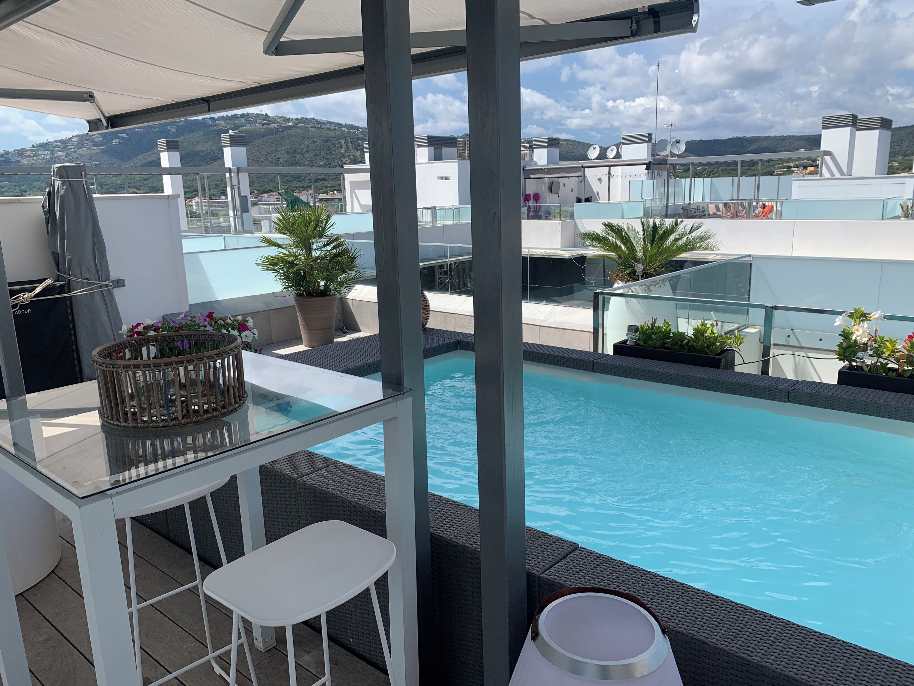 A rooftop pool area with a glass table and bar stools, surrounded by potted plants and overlooking a cityscape with hills.