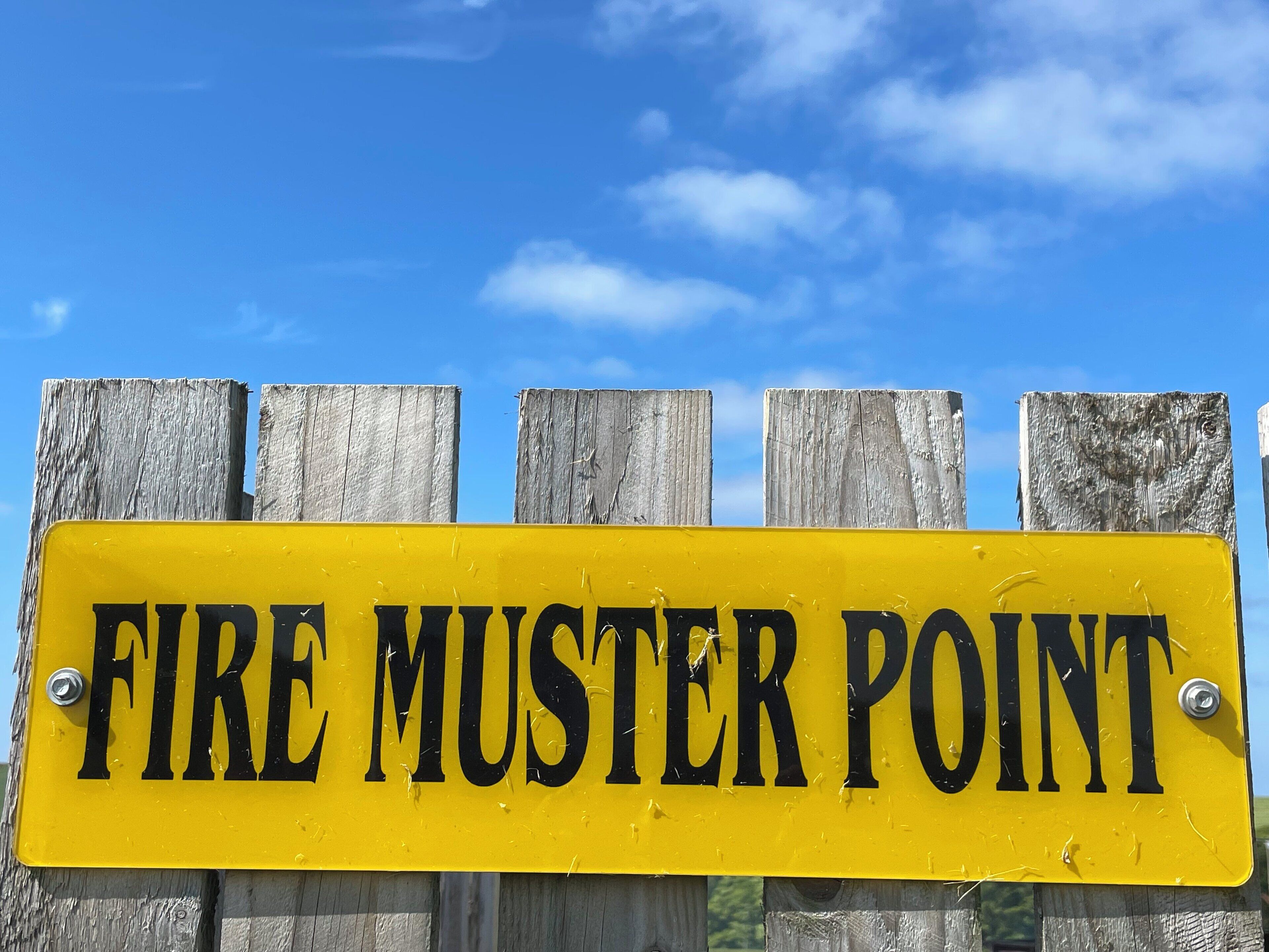 A yellow sign with black text reading "FIRE MUSTER POINT" mounted on a wooden fence.