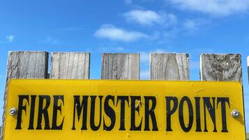 A yellow sign with black text reading "FIRE MUSTER POINT" mounted on a wooden fence.