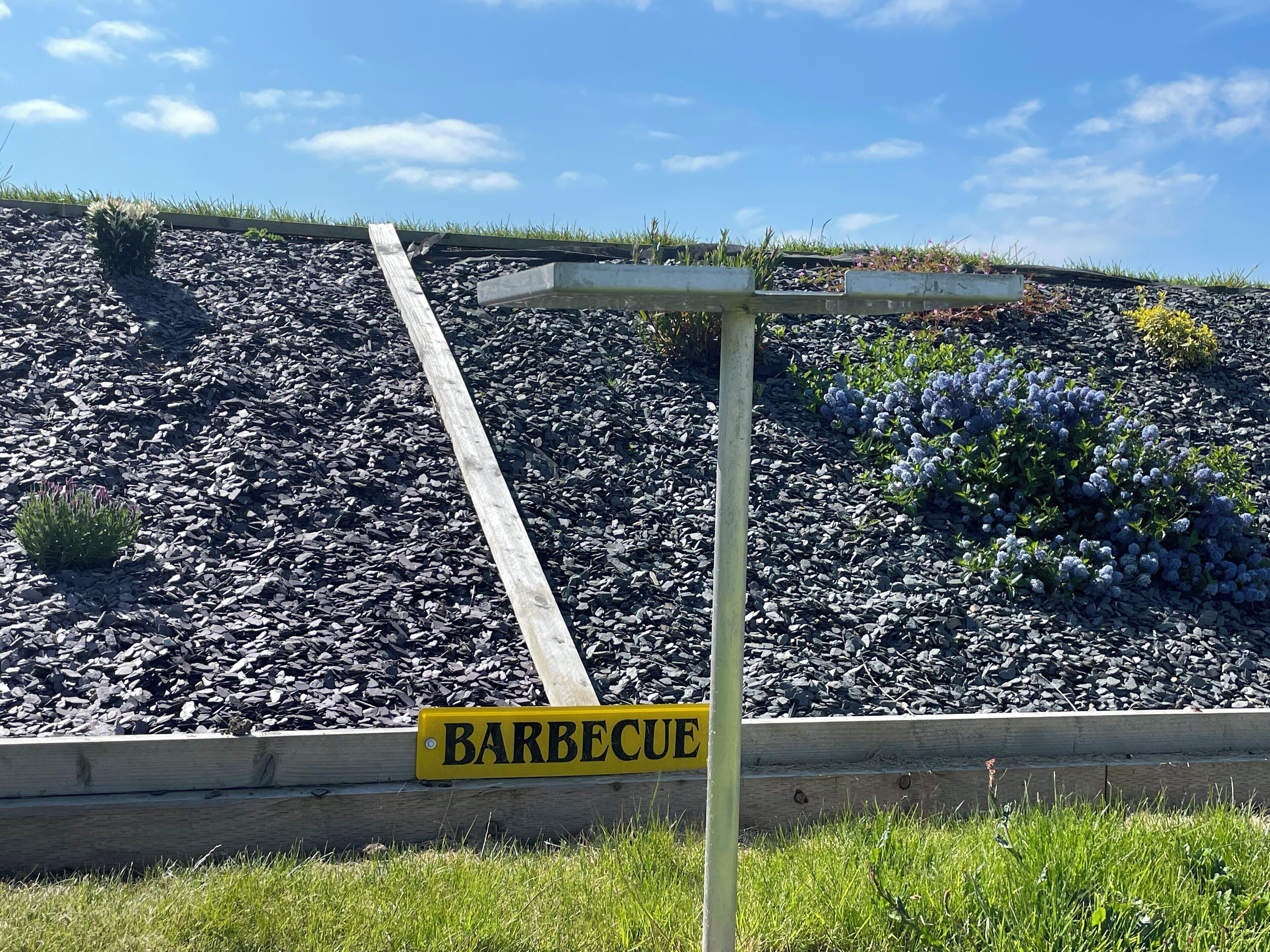 Schild mit der Aufschrift 'BARBECUE' vor einer Grünanlage mit einer Holzkonstruktion und einem mit Kies bedeckten Hang.