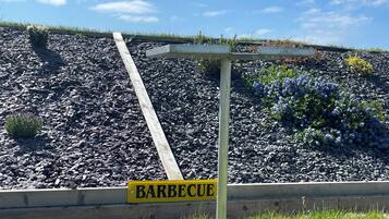 Schild mit der Aufschrift 'BARBECUE' vor einer Grünanlage mit einer Holzkonstruktion und einem mit Kies bedeckten Hang.