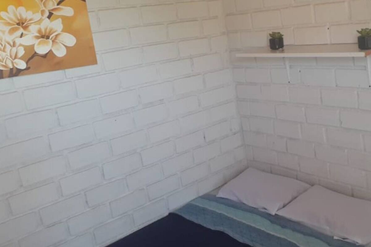 A small, white brick-walled room with a bed, pillows, a shelf with plants, and a painting of flowers.