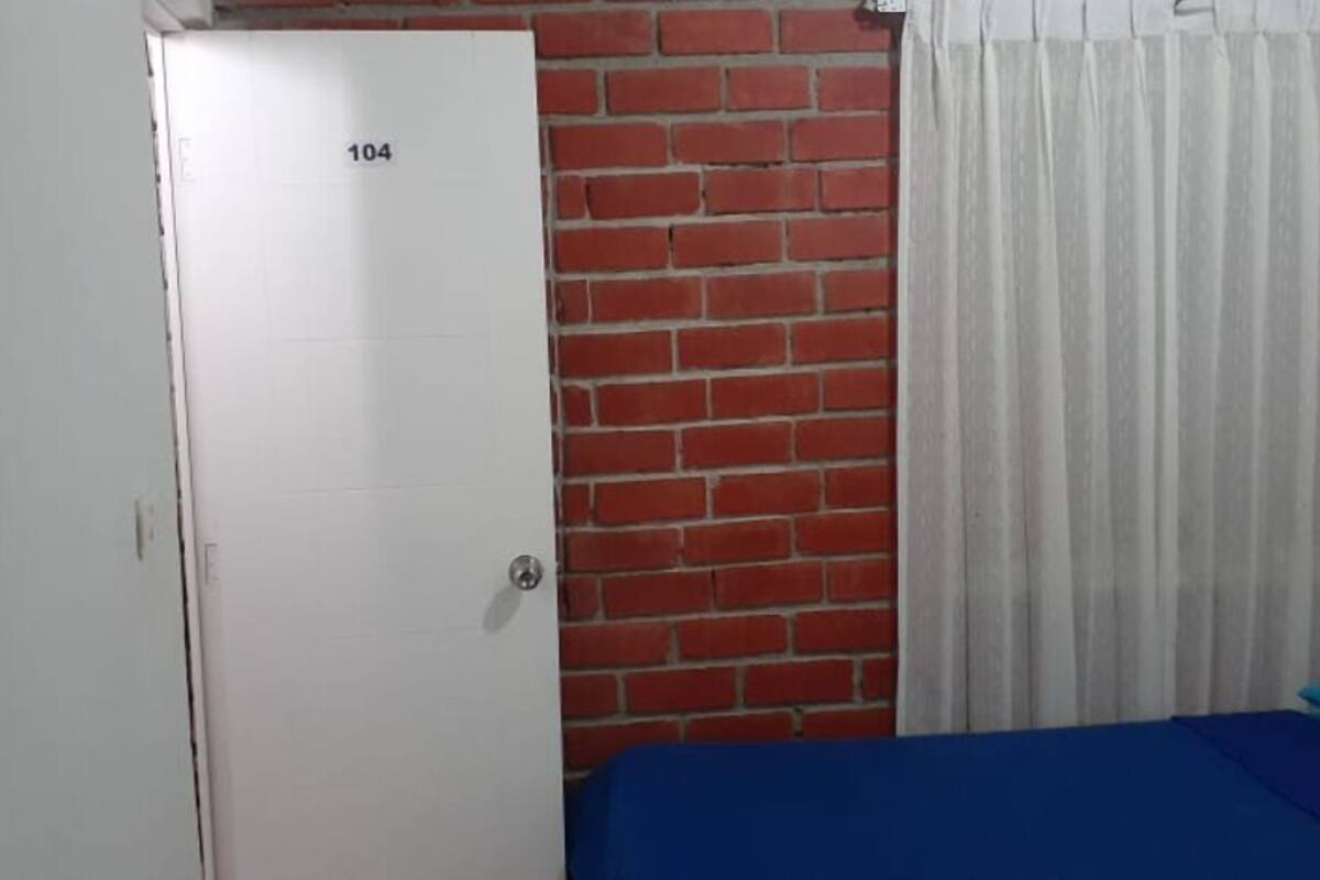 A room with a bed, a white door with the number 104, a brick wall, and white curtains.