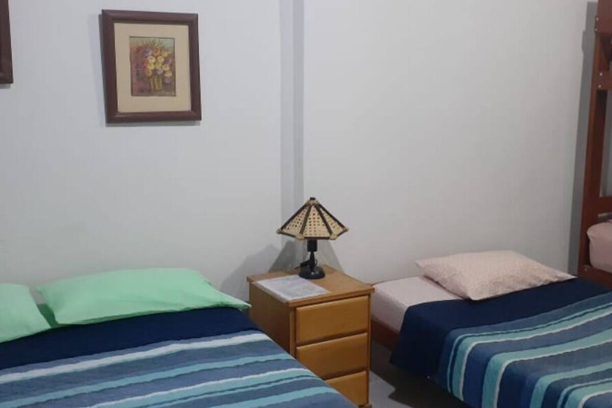 A dormitory room with three beds, each with different colored bedding. There is a nightstand with a lamp between the beds.