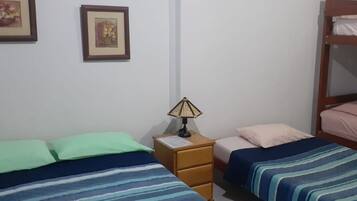 A dormitory room with three beds, each with different colored bedding. There is a nightstand with a lamp between the beds.
