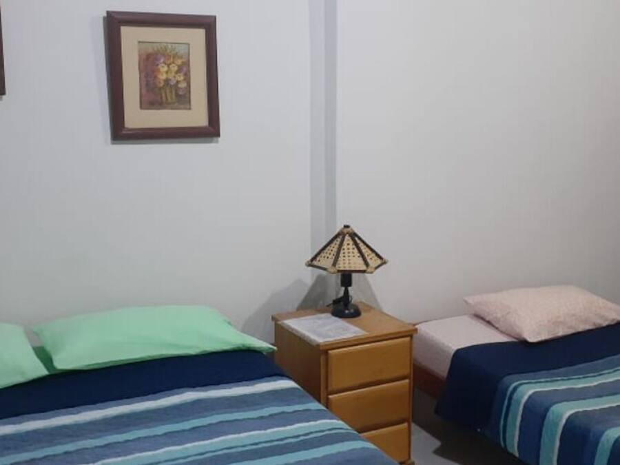 A dormitory room with three beds, each with different colored bedding. There is a nightstand with a lamp between the beds.