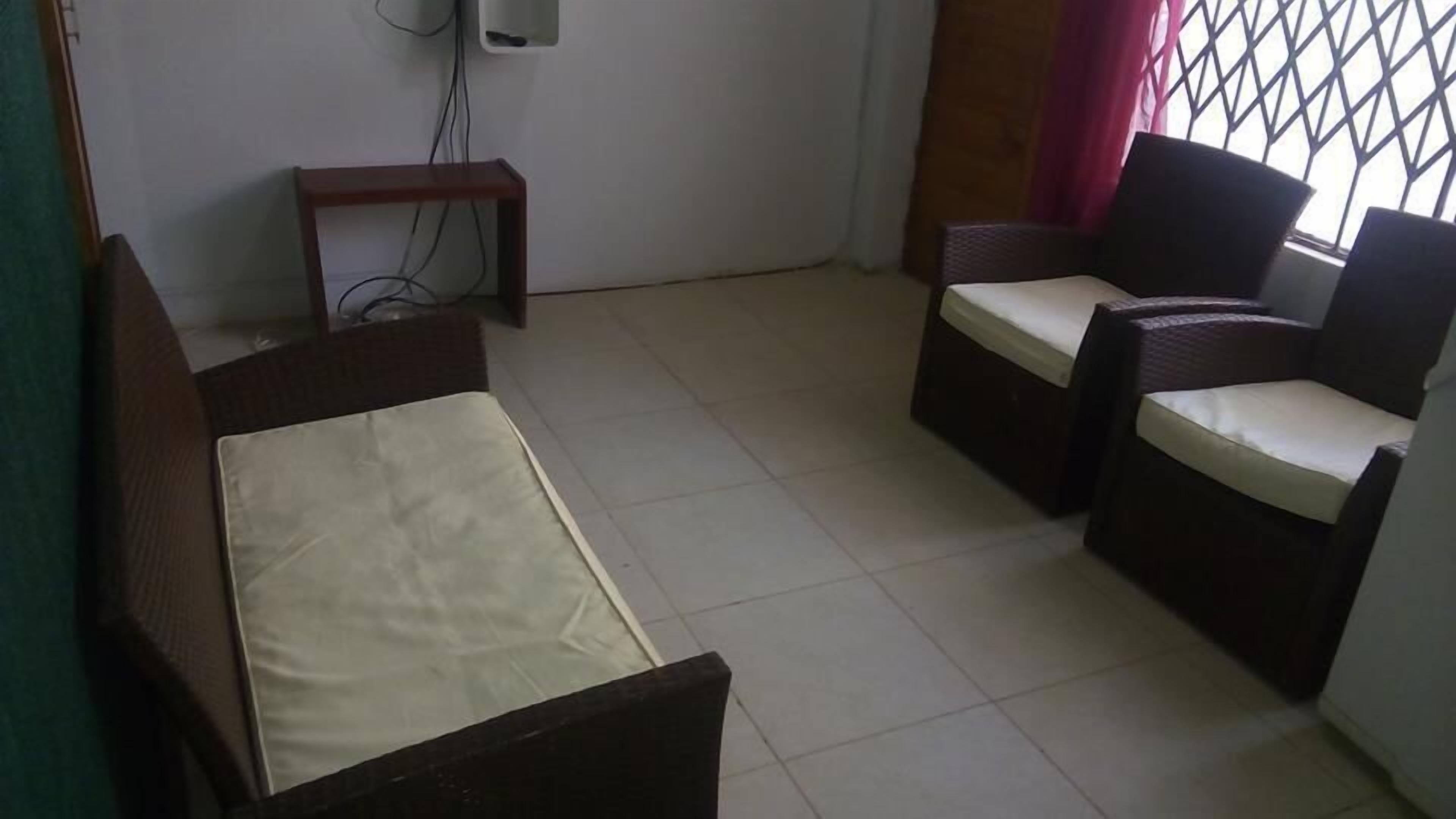 Una stanza piccola e arredata in modo essenziale, con un letto, due sedie, un tavolino e un altoparlante a parete.