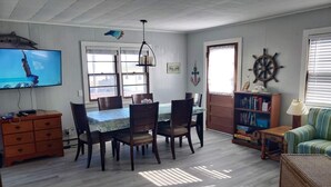 Un coin repas avec une table et des chaises, un téléviseur, une bibliothèque et une fenêtre avec des stores.