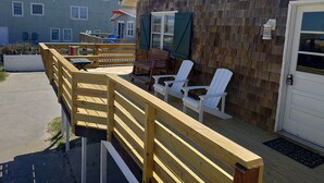 Une terrasse en bois avec des chaises blanches et une table, une porte avec une boîte aux lettres, et un bâtiment dont l’extérieur est recouvert de bardeaux.