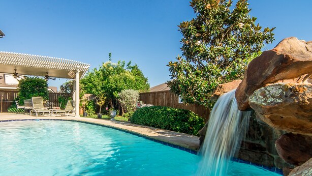 A swimming pool with a waterfall feature, surrounded by a wooden deck and outdoor furniture. - 4 Bedroom With Landscaped Private Pool & Outdoor Pool Table ~MONTHLY DISCOUNT~ (McKinney)