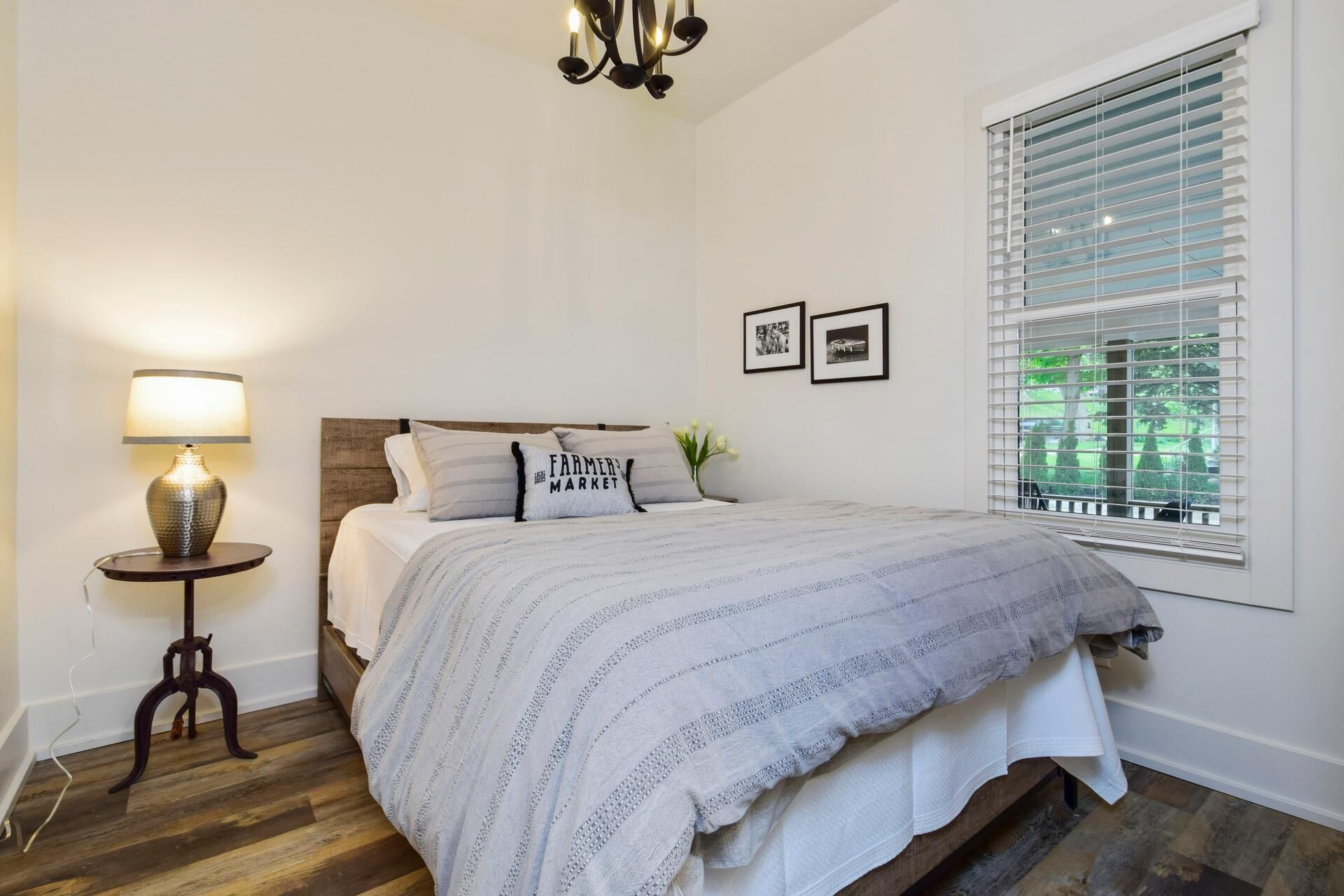 A bedroom with a bed, a bedside table with a lamp, a window with blinds, and two framed pictures on the wall.