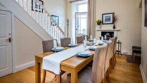Uma sala de jantar com mesa de madeira posta para seis lugares, ladeada por cadeiras, uma escadaria e uma lareira.