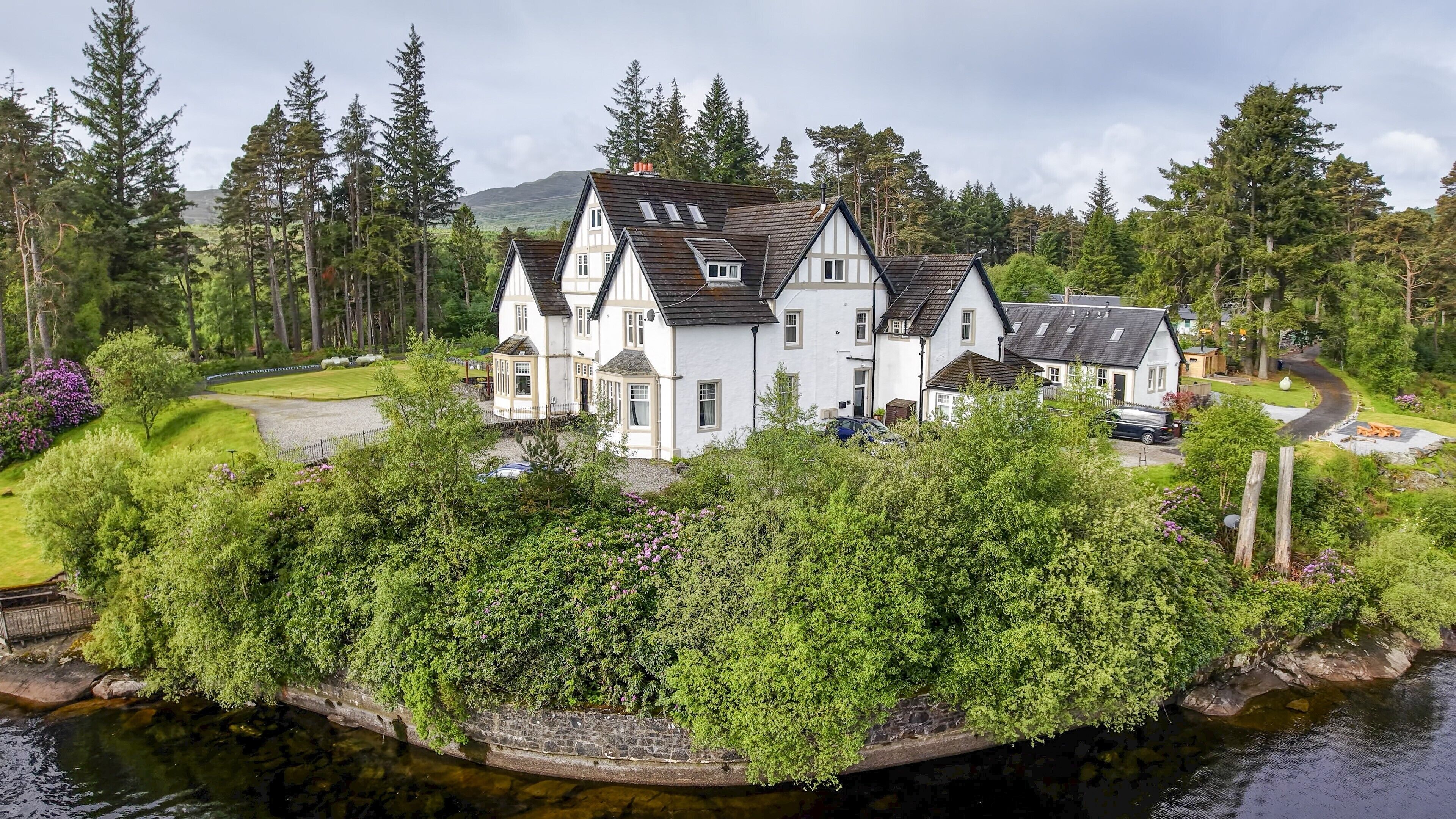 A large white house with multiple chimneys, surrounded by lush greenery and a body of water.