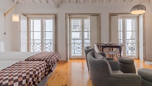 A spacious room with a wooden floor, a checkered bed, grey upholstered chairs, a dining table, and large windows with a balcony.