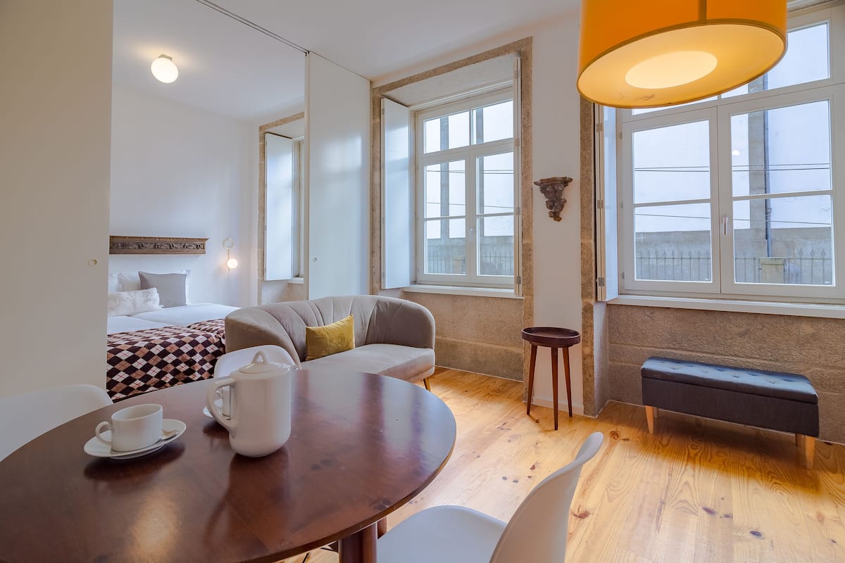 Habitación de hotel con cama, sofá, mesa de comedor con tetera y taza, banco y ventanas amplias con vistas.