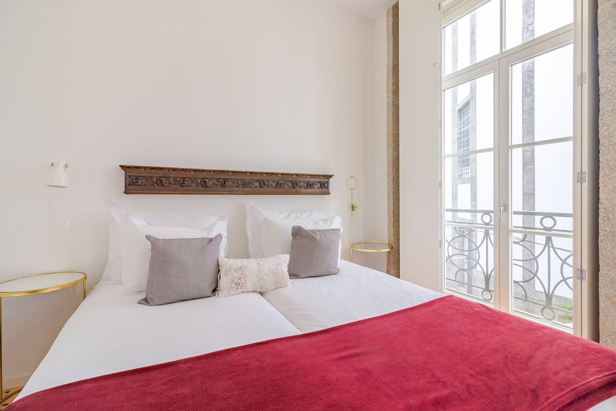 Una cama bien tendida con sábanas blancas y una colcha roja, un cabecero decorativo y vistas a un balcón con barandillas de metal.