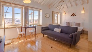 A modern room with a wooden floor, a grey sofa, a dining table with chairs, and large windows with a view of the city.