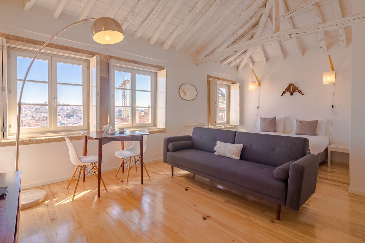 Una habitación moderna con suelo de madera, un sofá gris, una mesa de comedor con sillas y ventanas amplias con vistas a la ciudad.