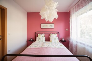 Ein Schlafzimmer mit einem Bett, einer dekorativen Deckenleuchte, einem gerahmten Bild an der Wand und einem Fenster mit transparenten Vorhängen.