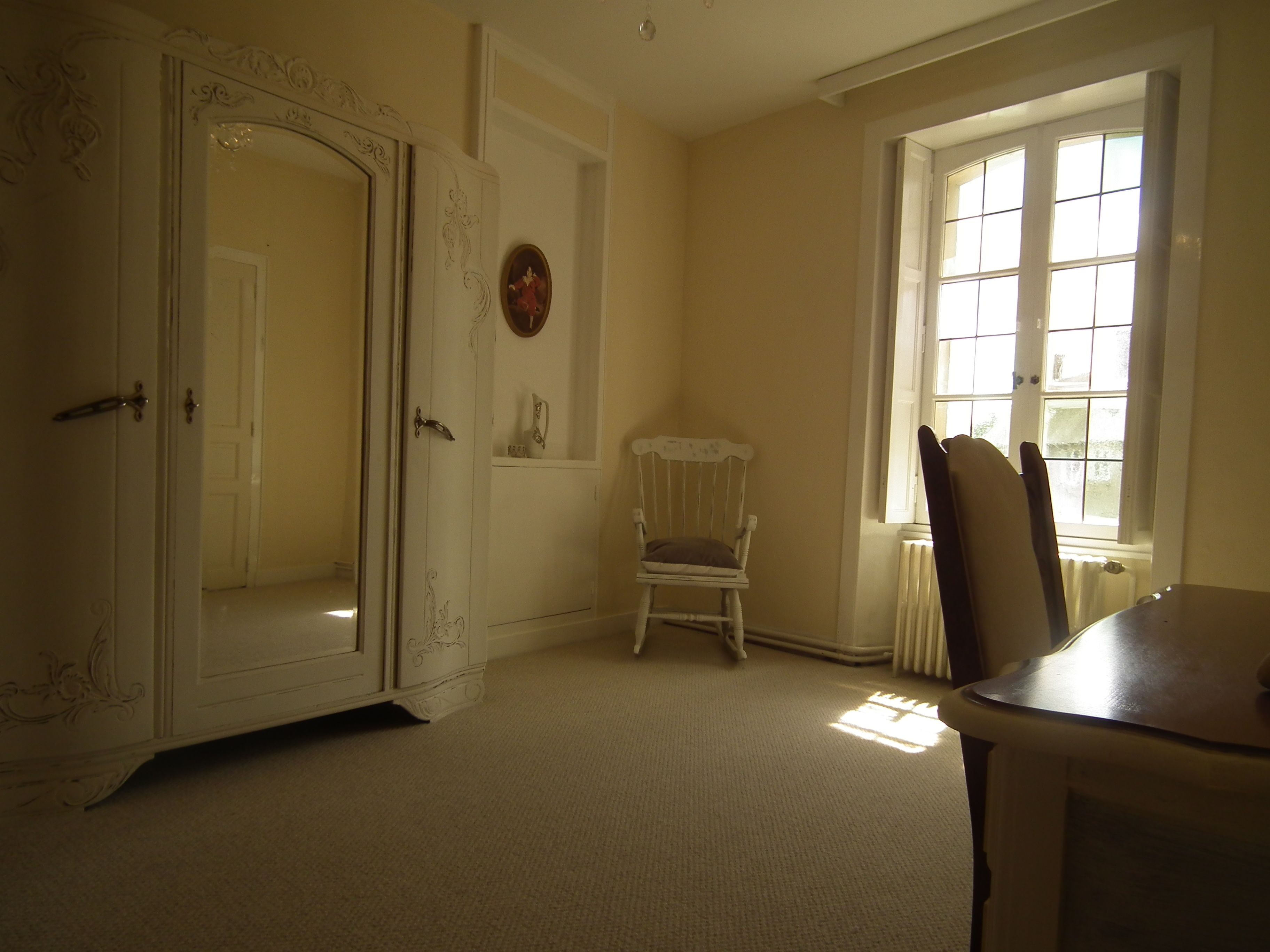 A room with a white wardrobe, a wooden chair, a table, and a window with natural light.