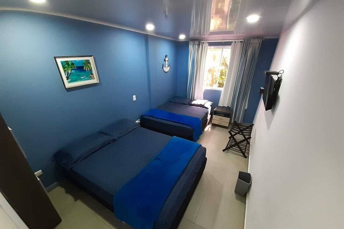 A room with two beds, a small table, a chair, a picture on the wall, and a window with curtains.