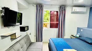 A modern hotel room with a grey sofa, a bed with a blue bedspread, a flat-screen TV mounted on the wall, and a window with striped curtains. - Palmare House (San Andrés)