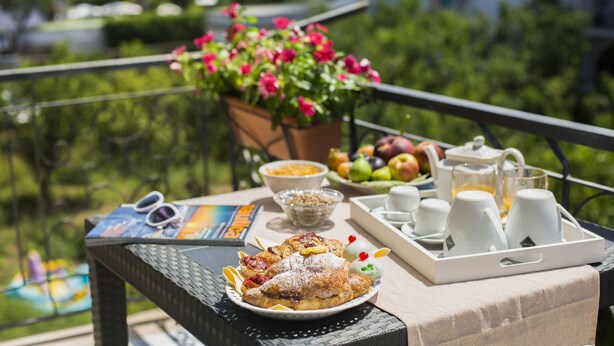 A breakfast setup on a balcony with a table, a tray of pastries, a bowl of cereal, a fruit bowl, and a coffee setup.
