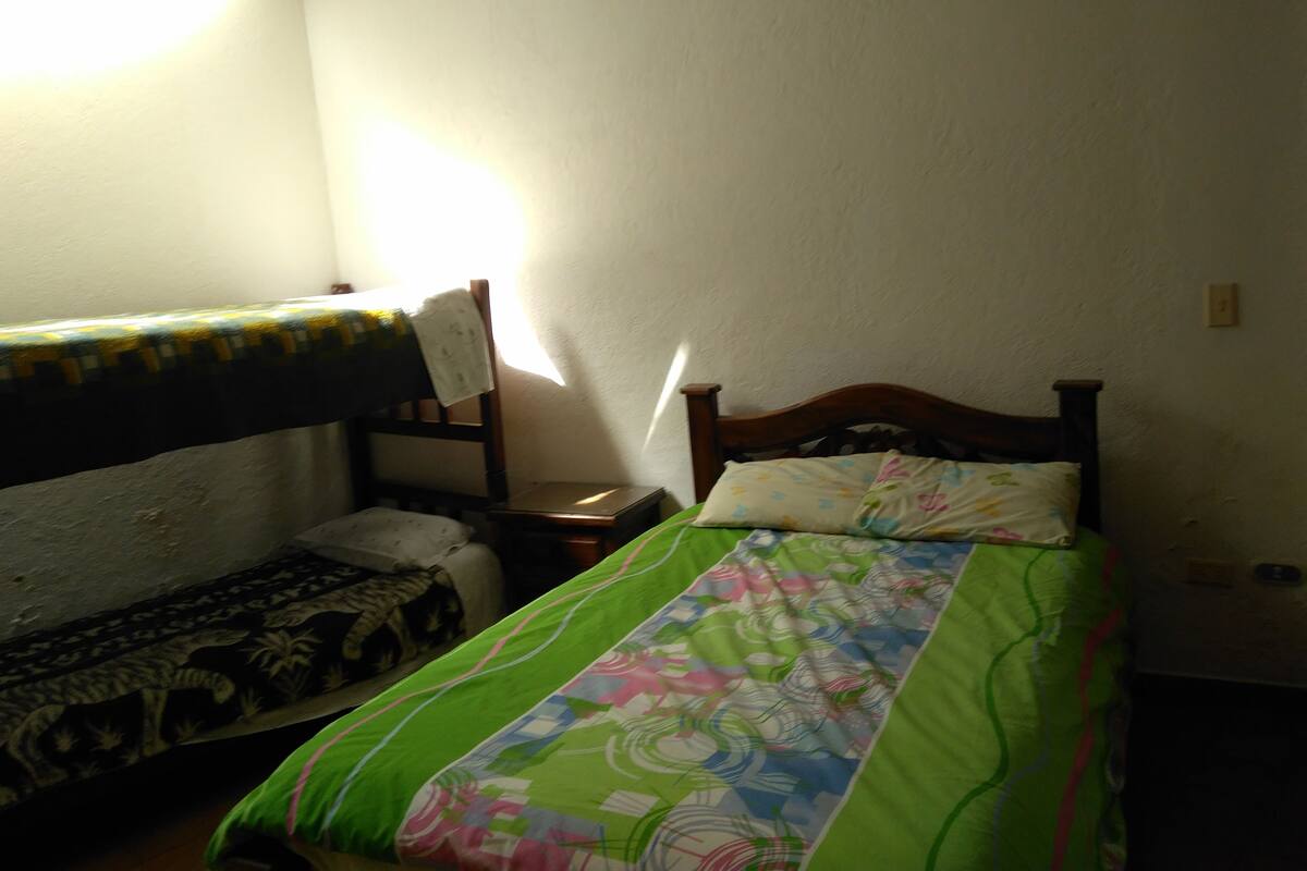 Ein kleiner, schwach beleuchteter Raum mit zwei Betten, eines mit einem gemusterten Überwurf und das andere mit einem bunten. Ein Stuhl und ein kleiner Tisch mit einer Lampe stehen ebenfalls im Raum.