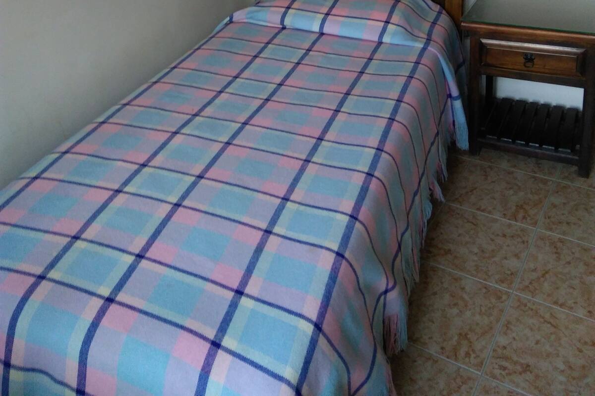 Ein Einzelbett mit Holz-Kopfteil und einer Karo-Bettdecke. Ein kleiner Nachttisch steht neben dem Bett.
