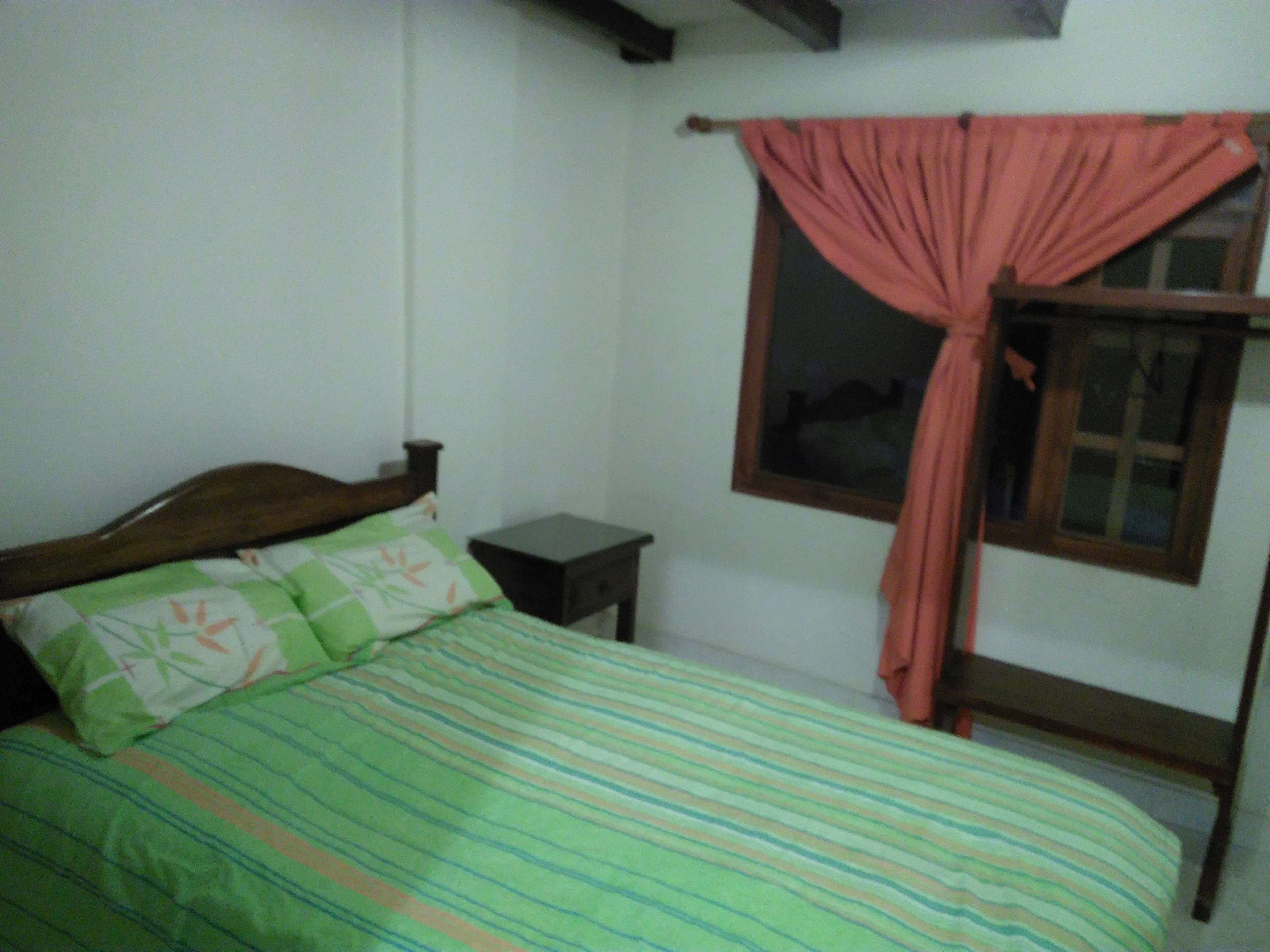 Habitación individual con una cama y cabecera de madera, una mesita de noche pequeña, una ventana con cortina y una pared lisa.