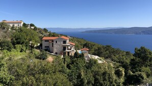Une propriété située sur la côte, avec une maison en pierre et un bâtiment  à toit rouge, entourée d’une végétation luxuriante et offrant une vue sur une mer bleue et limpide.
