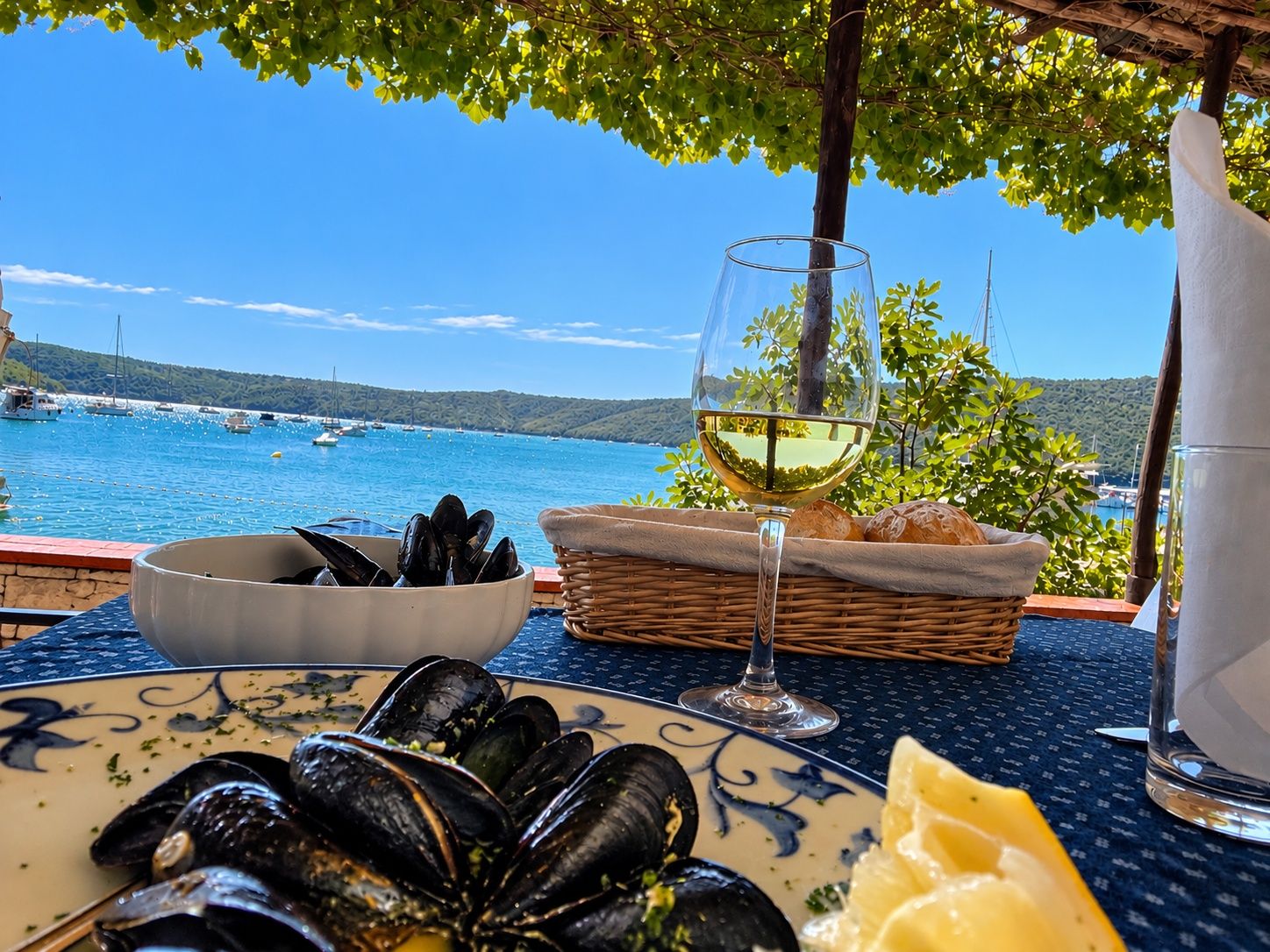 Un tavolo con cozze, un bicchiere di vino bianco, pane e spicchio di limone, con vista su un porto turistico e barche.