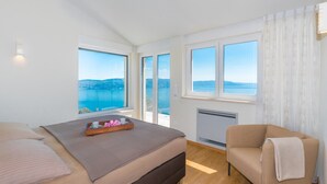 Une chambre à coucher avec un lit, une chaise et une vue sur une étendue d’eau, visible à travers de grandes fenêtres.