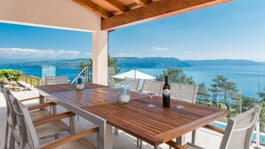 Une table en bois avec des chaises, une bouteille de vin et un verre, sur une terrasse offrant une vue sur la mer et les montagnes.